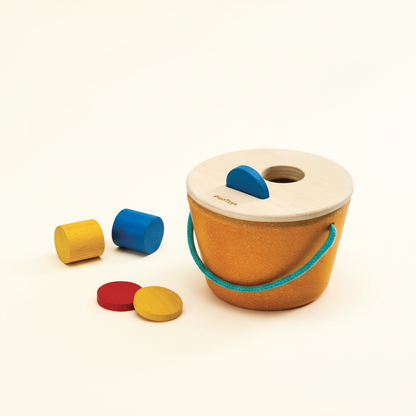 PlanToys wooden bucket with lid and shaped wooden pieces on a plain cream background